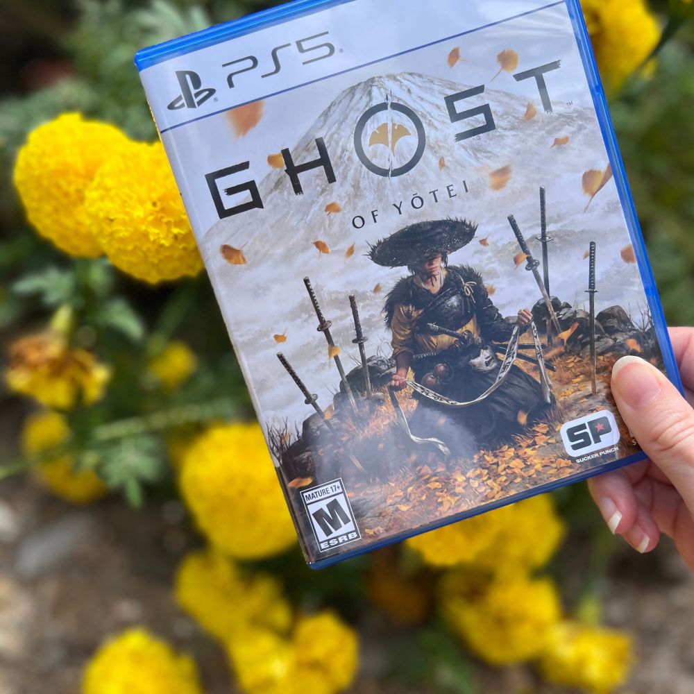 A physical copy of Ghost of Yōtei for PlayStation 5 held in front of yellow flowers