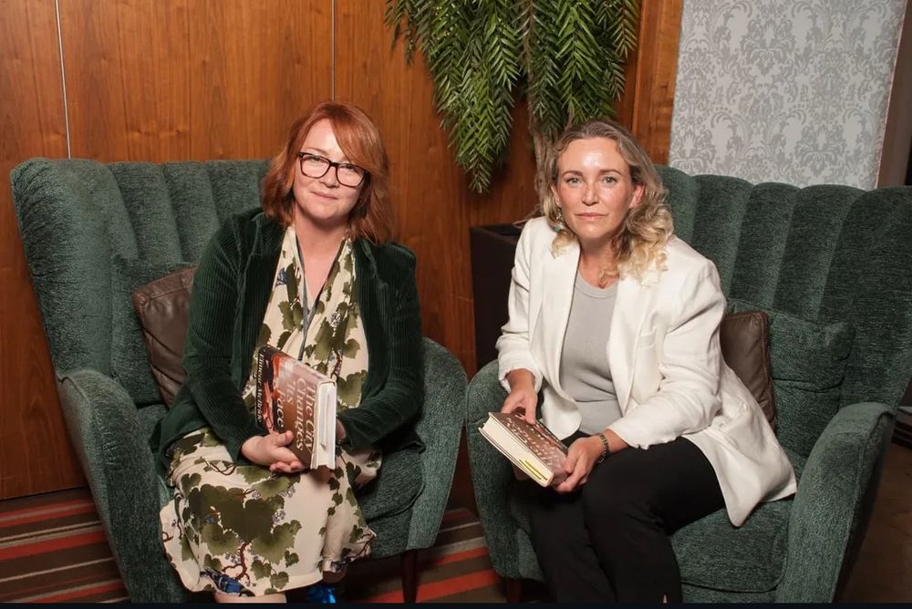 Eimear McBride and Cristín Leach sitting next to each other in dark green upholstered armchairs, each holding a hardback copy of Eimear’s new novel, The City Changes Its Face. 