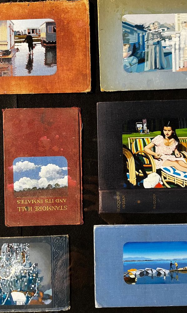 Selection of Catherine Barron book cover paintings - images painted on vintage hardback linen covered book covers. These are displayed in table top vitrines in the gallery. This is a shot taken through the vitrine glass looking down, showing some painted covers. Sky, lake, a woman sitting on a sun lounger. 