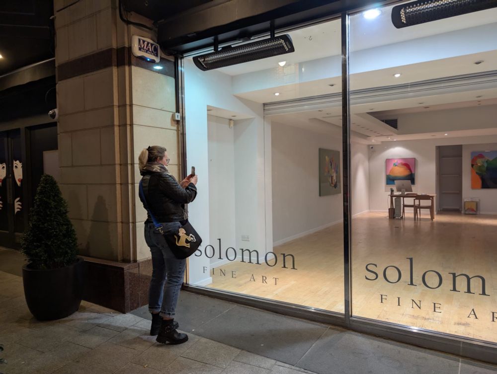 A woman in jeans and black leather jacket takes a pic with her phone through the full-length glass windows of the Solomon Gallery in Dublin at night. Inside, colourful paintings by Tom Climent are hung on the walls. 