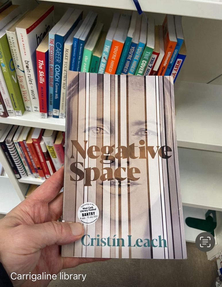 A hand holding a copy of my memoir Negative Space in front of a set of white bookshelves half filled with other books behind. 

The cover of my book features a glass plate sepia hued photograph of my face taken by Alex Sapienza in Dublin in 2012, overlaid with vertical white/sepia stripes which semi-obliterate the close up like vertical blinds. My eyes are most visible. The words negative space cross my nose and top of mouth. My name Cristín Leach is in green at the bottom of the cover. This cover has a sticker which reads Appearing at the West Cork Literary Festival Bantry 8 July - 15 July 2022. 

Cover design is by Fiachra McCarthy.