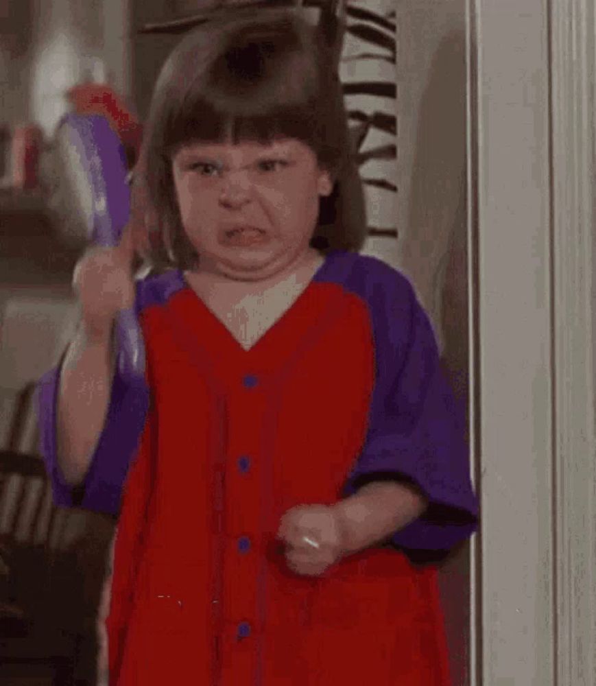 a little girl in a red and purple dress is making a funny face while standing in front of a door .