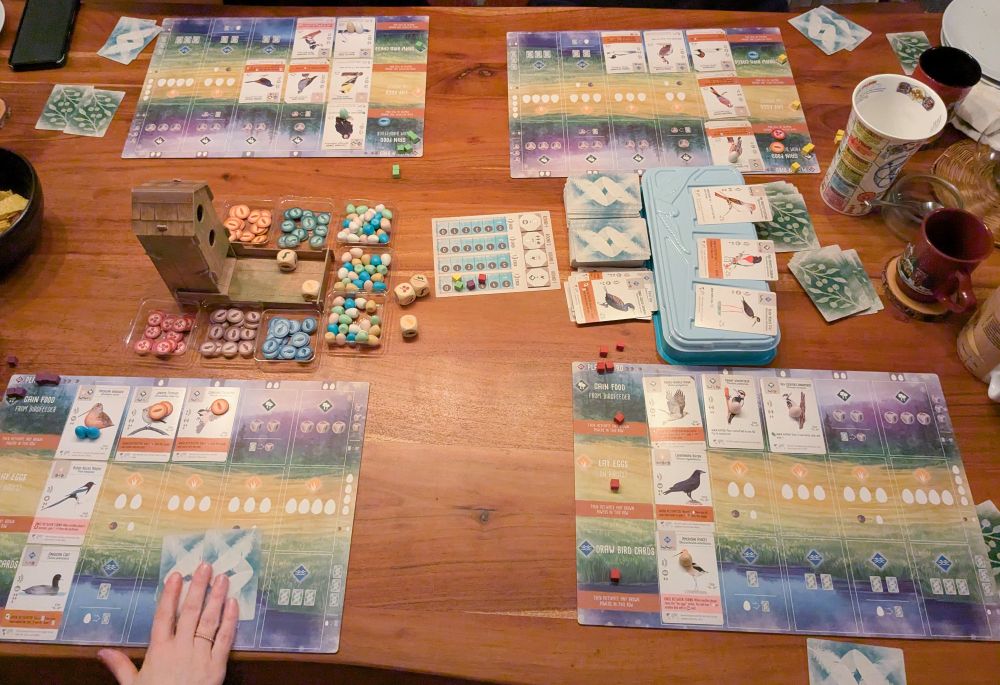 A photo of all our Wingspan game mats out on a table. There's also a bird feeder thing in the middle (part of the food in the game), some stacks of cards, and some mugs for the mandatory hot drinks