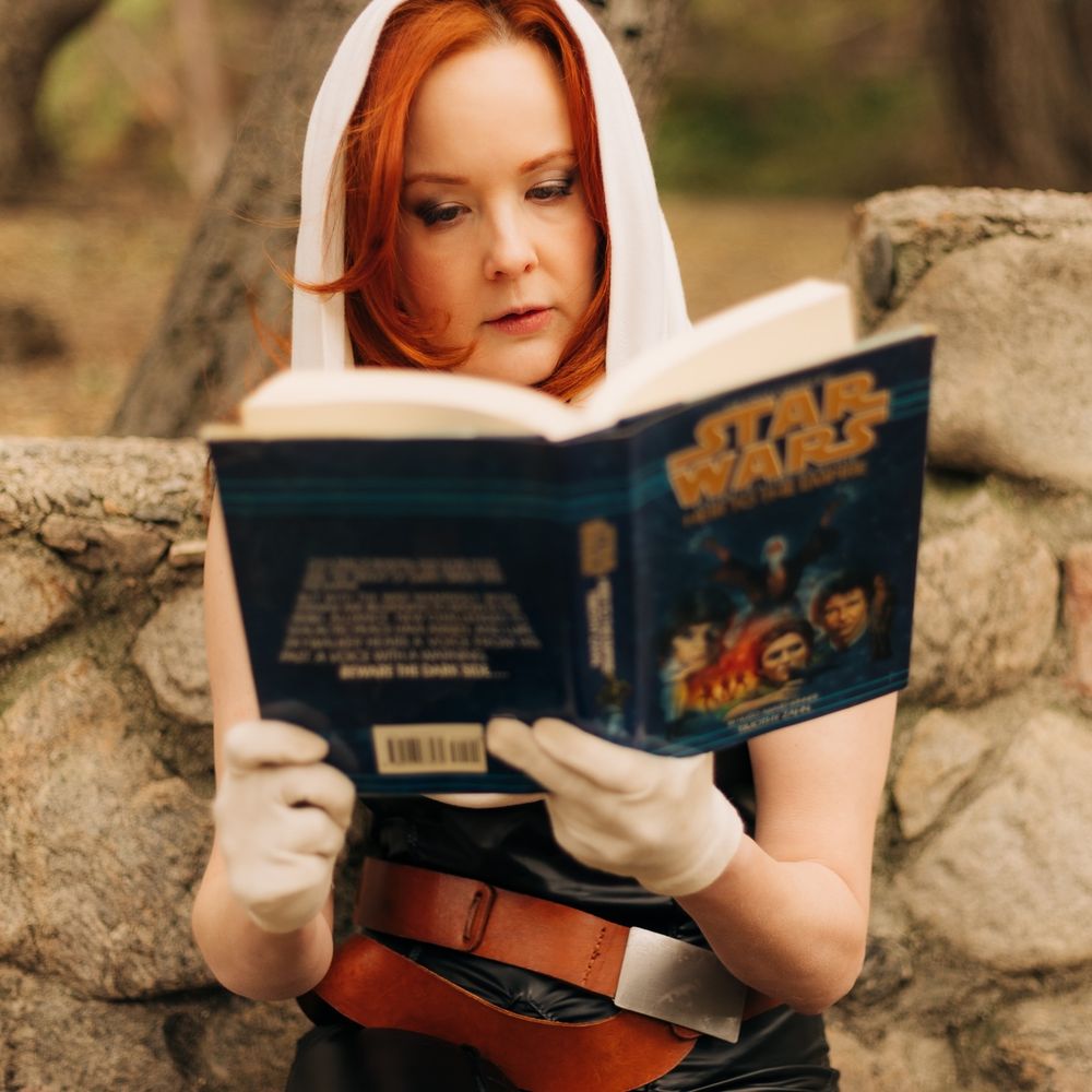 A Mara Jade cosplayer holds an open copy of Heir to the Empire in front of her face, reading.