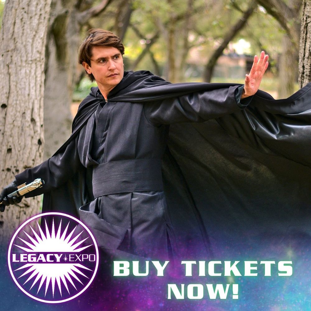 A photo of an episode 6 Luke Skywalker cosplayer. He is holding his lightsaber hilt in one hand and is throwing his other up as though pushing something with the Force. His black cape is tossed up dramatically. 
