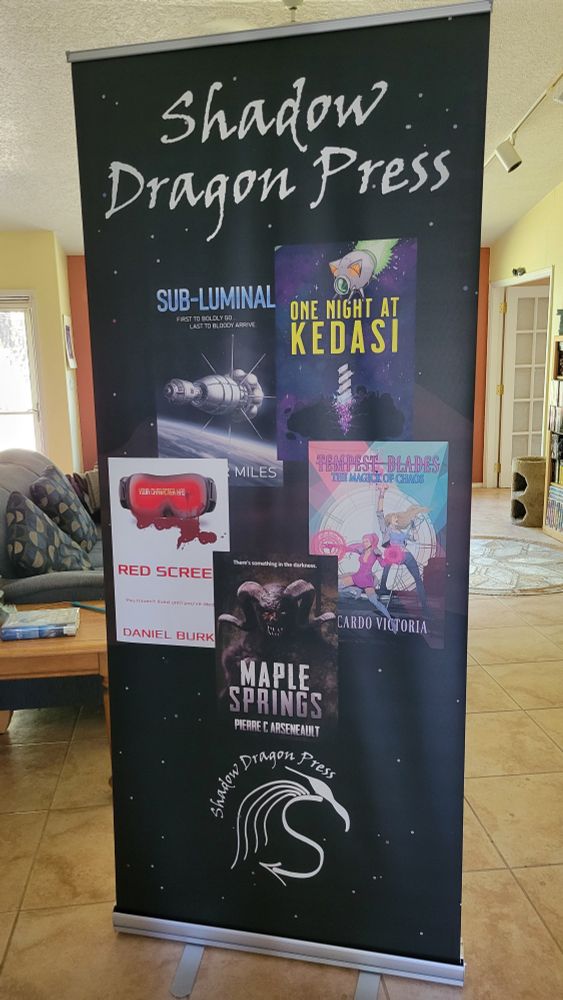 Large stand up banner with "Shadow Dragon Press" at the top amd a collection of five book cover images beneath. Books featured are Sub-Luminal, One Night at Kedasi, Red Screen, The Magick of Chaos, and Maple Springs.