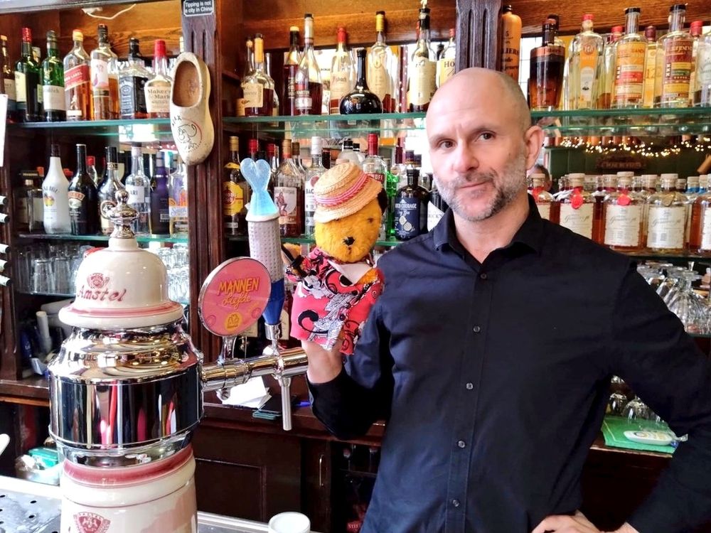 The barman with our Sooty puppet behind his bar at Louis Bar in Amsterdam. The shelves behind them are full of bottles & a wooden clog is hanging on a hook