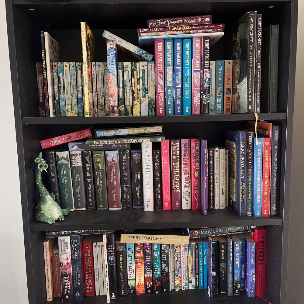 A photo of a bookshelf filled with Terry Pratchett novels in various editions, so full some books lie on their side on top of the others. A friendly green dragon statue with a long neck sits at the left end the middle shelf.