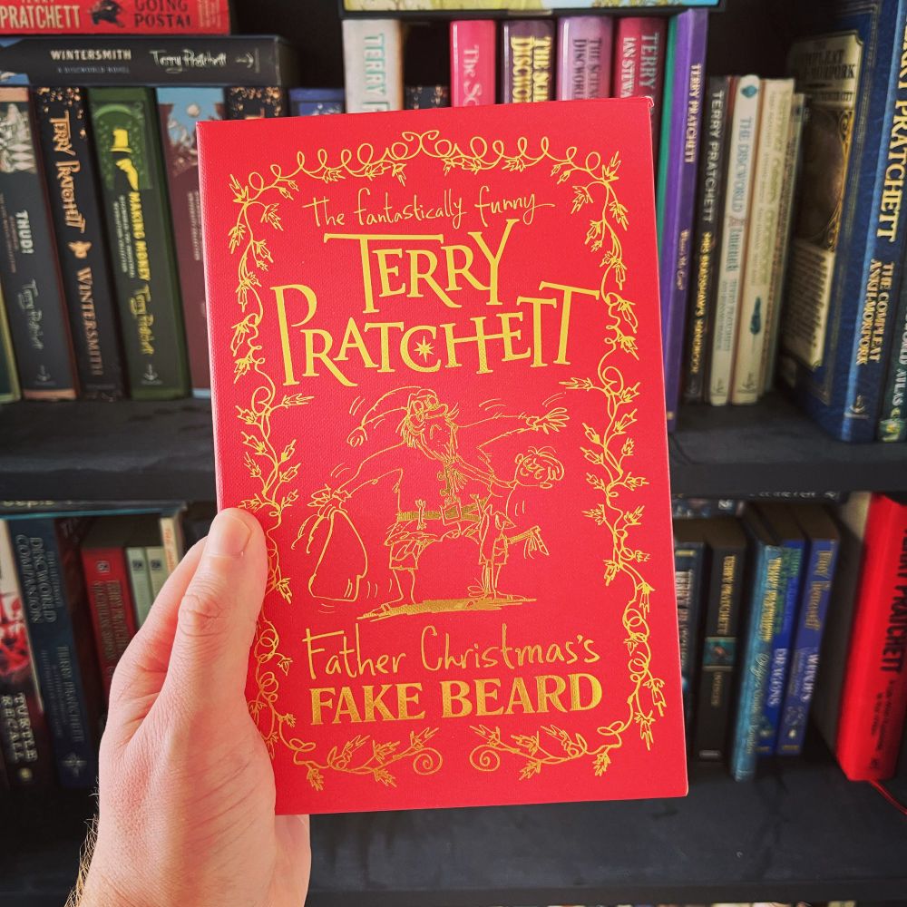 A photo of the deluxe hardcover edition of Father Christmas’s Fake Beard by Terry Pratchett, held in front of a bookshelf full of other Terry Pratchett books.