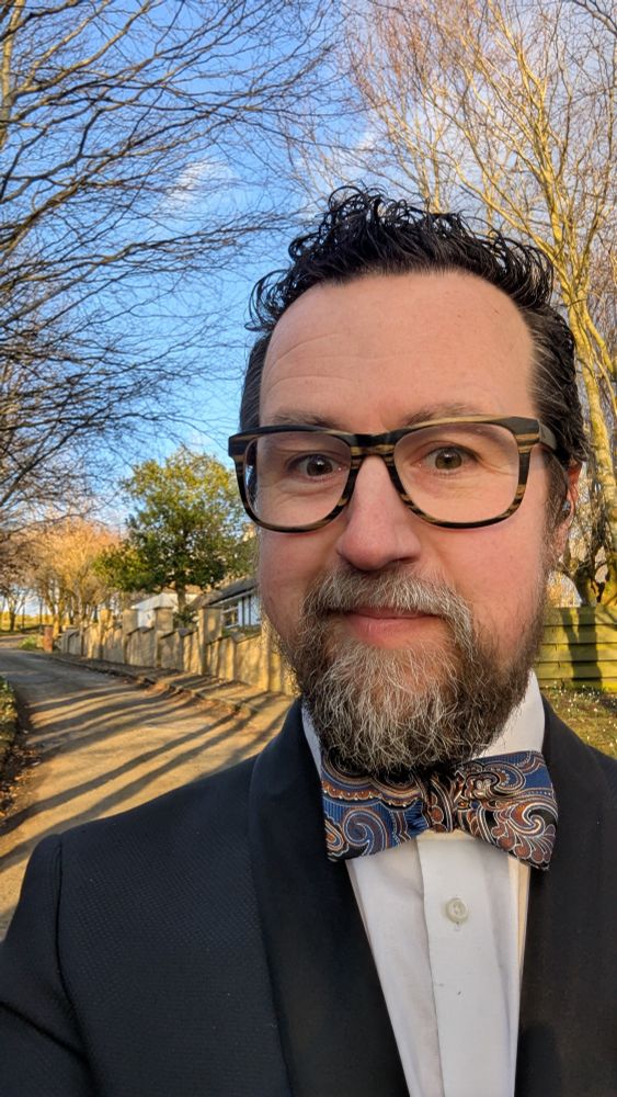 Josiah wearing a tuxedo with a paisley bow tie. Standing outside in the forest.