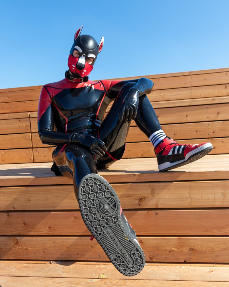 Man in a black and red full body rubber suit with dog mask and red, black and white adidas shoes. Focus on the sneakers. Sitting on wooden stairs/bench