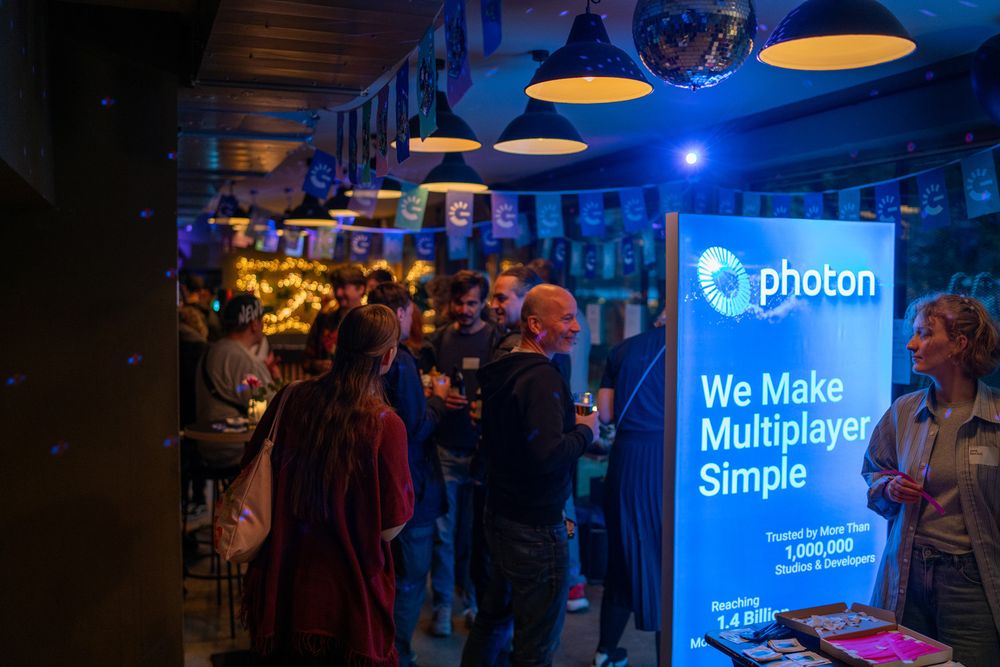 “Series of photos showing a vibrant networking event held indoors and outdoors under warm, festive lighting. The venue features large canopies, hanging lamps, and decorative string lights, with colorful banners displaying a hand-shaped logo. Attendees are gathered in groups, chatting and holding drinks around high tables and bar counters. Some images show structured activities with people seated at long wooden tables, engaged in conversation, with drinks and small flower arrangements on the tables. A prominent illuminated sign in one photo reads ‘Photon – We Make Multiplayer Simple,’ highlighting a sponsor presence. The overall atmosphere is casual and lively, with a mix of standing and seated interactions in a cozy, modern setting.”