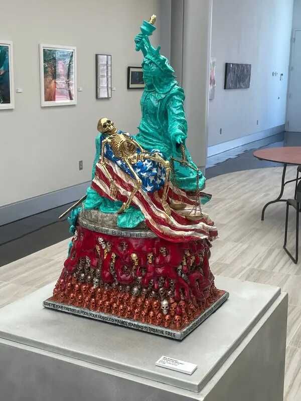 A sculpture on a pedestal in a museum. The base of the sculpture is a pile of red or gold human skills. Atop the pile, the Statue of Liberty is bent over, staring a gold human skeleton lying on an American flag.