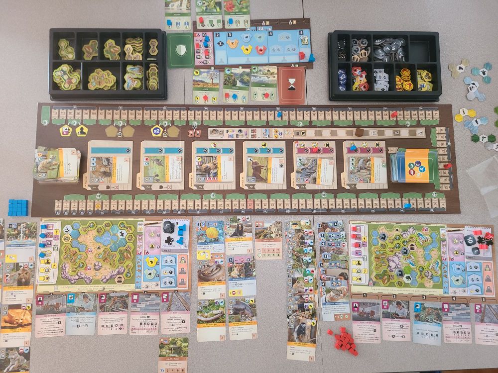 The board/table for a finished game of Ark Nova. Several animal, sponsor, research, and objective cards, and blue and red pieces for each player, are strewn about the table.