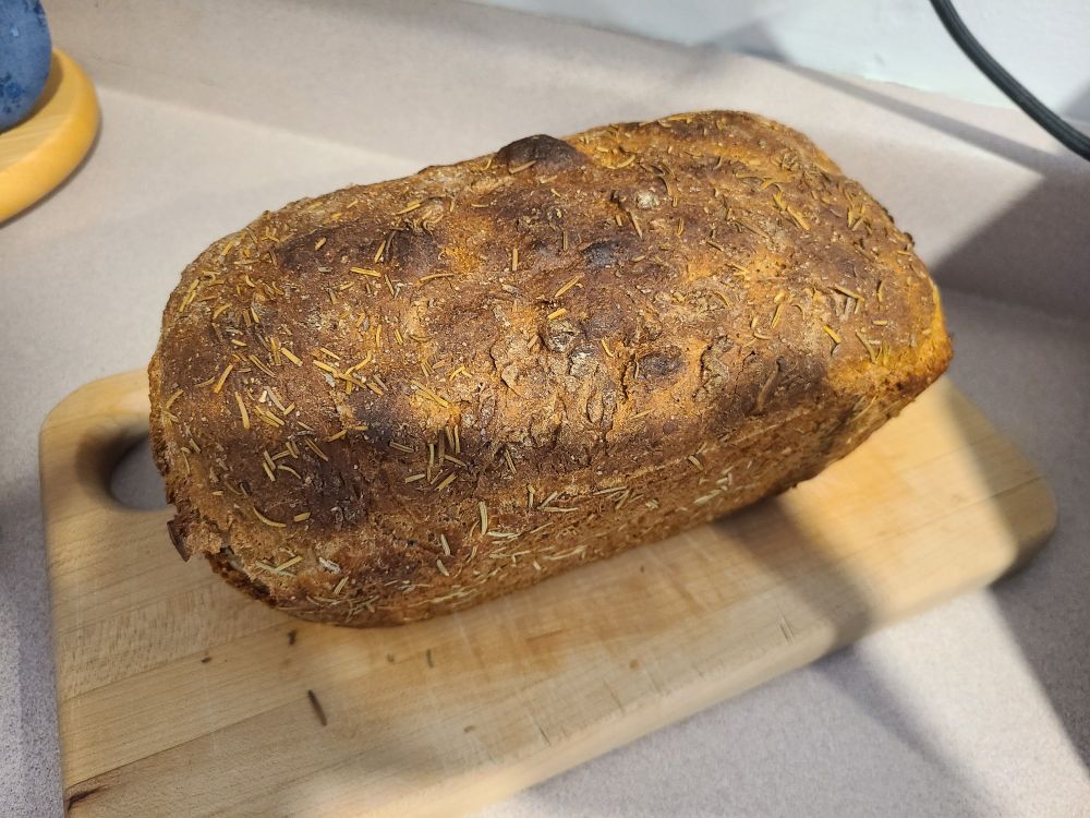 A whole loaf of bread made with chickpea flour, seasoned with rosemary, sits on top of a wooden cutting board.
