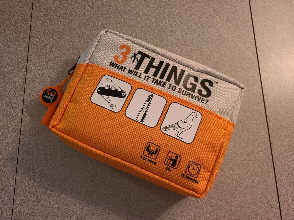 The fabric case for 3 Things, with attached whistle. The front says, "What will it take to survive?", with icons of a multitool, lipstick, and a pigeon.