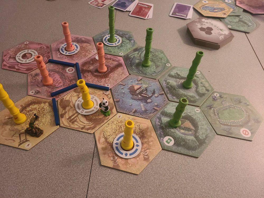 Board at the end of a game of Takenoko. Several multicoloured land hex tiles are host to growing columns of bamboo. A gardener and a panda stand on two of the tiles.
