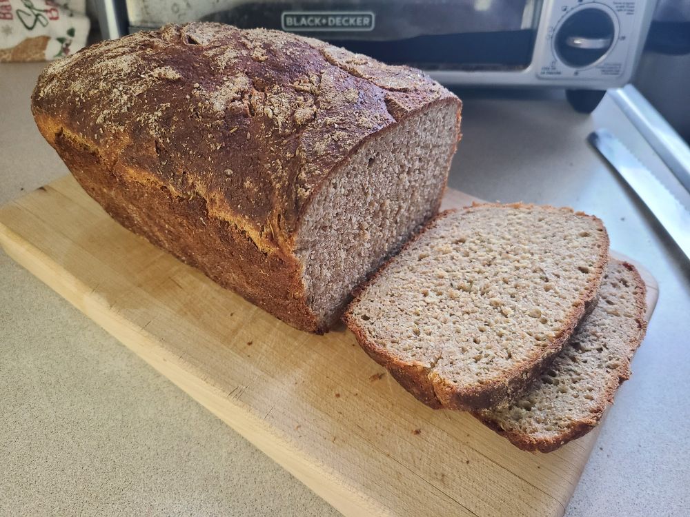 A loaf of light brown bread with a fine, fairly uniform crumb and a darker brown, textured crust, lies on a cutting board with two slices taken out of it.
