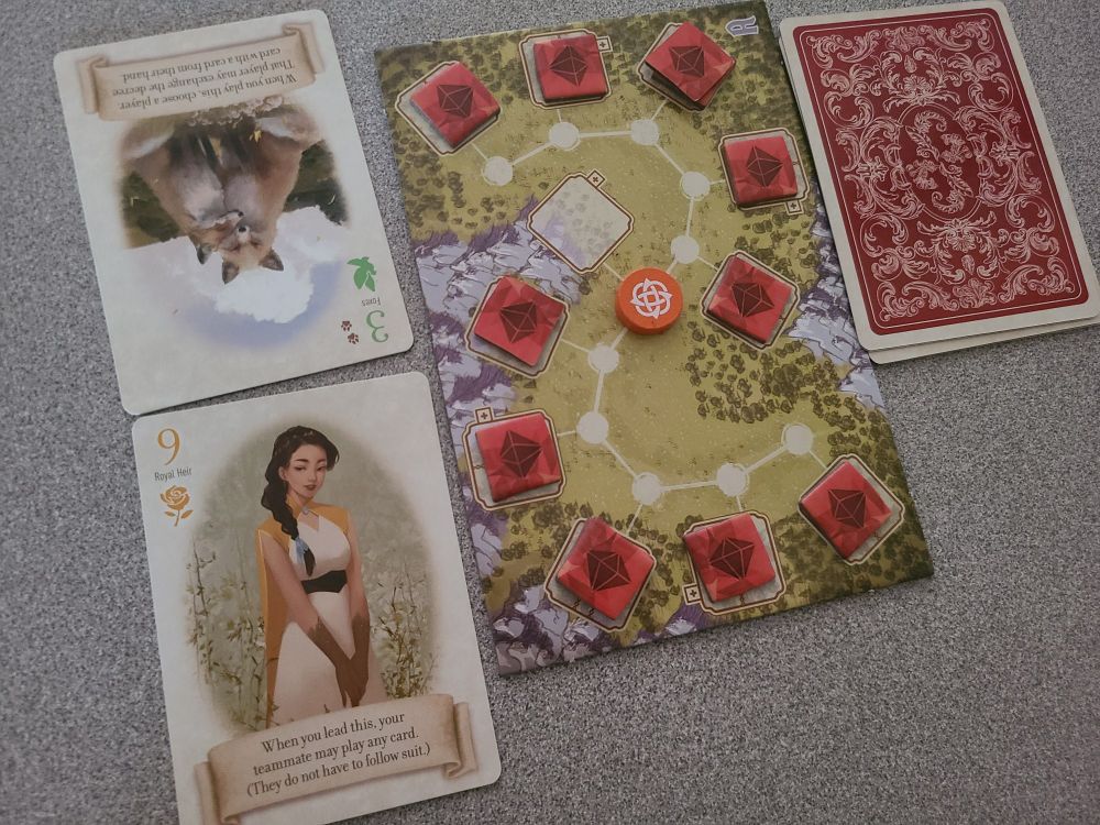 A sample game of The Fox in the Forest: Duet. There is a board with an S-shaped path and a player token in the middle space of that path, and several red gem tokens attached to the other spaces on that path. Two cards from the deck are laid out on one side, the 3 (Foxes) of Doves and the 9 (Royal Heir) of Roses, each with associated abilities printed on the cards.