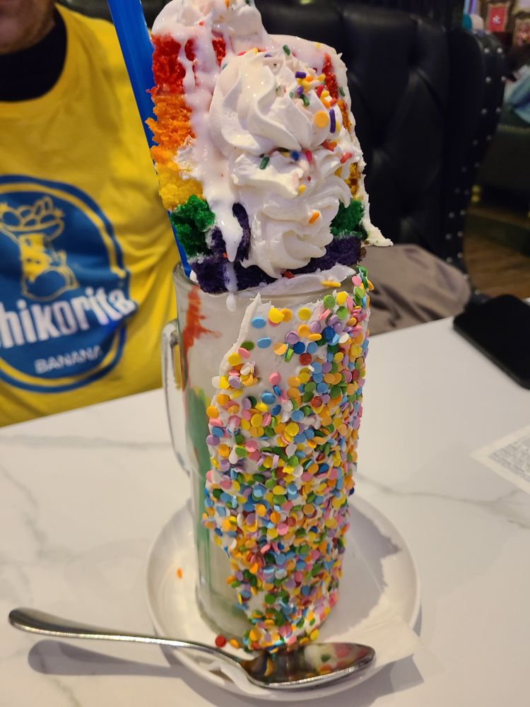An abomination of a rainbow sprinkled milkshake with an entire slice of rainbow cake on top.