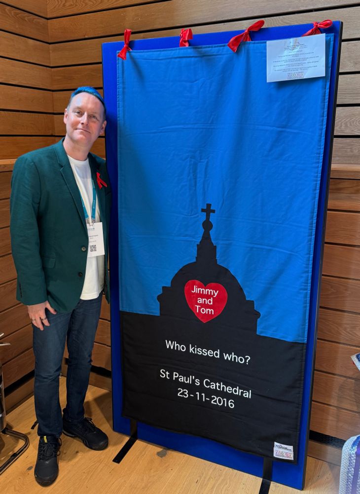 Tom, wearing a teal jacket and jeans, stands in front of his HIV living quilt. (Described in next image)
