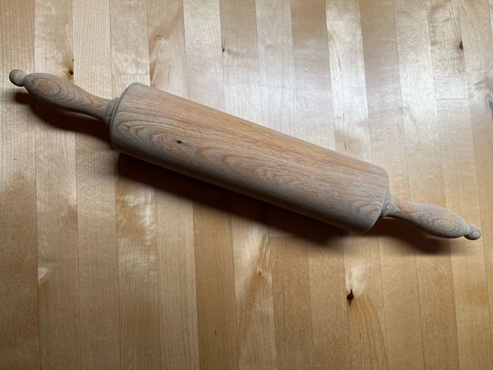 Ein Nudelholz aus Holz mit geschwungenen Griffen liegt auf einem Holztisch
