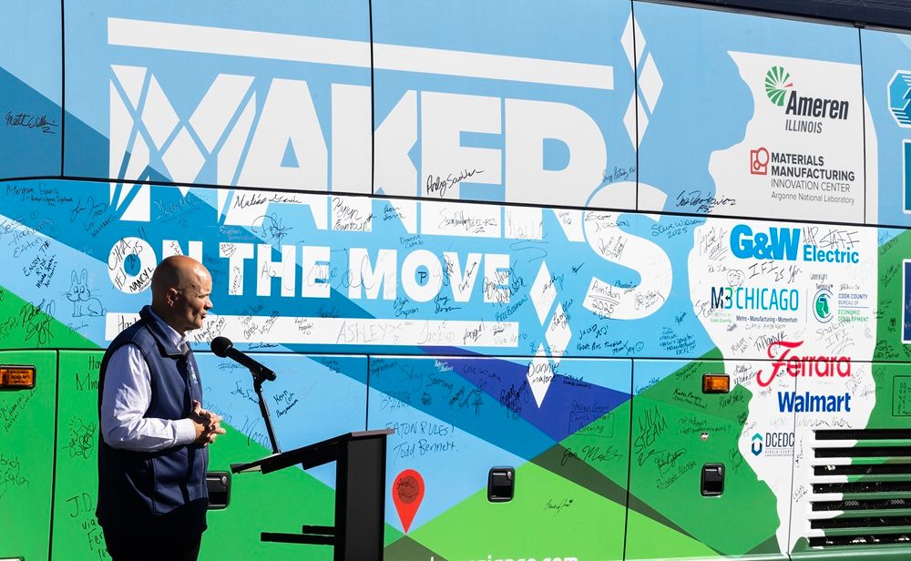 Man speaks at a podium in front of the Makers on the Move bus covered with signatures and logos