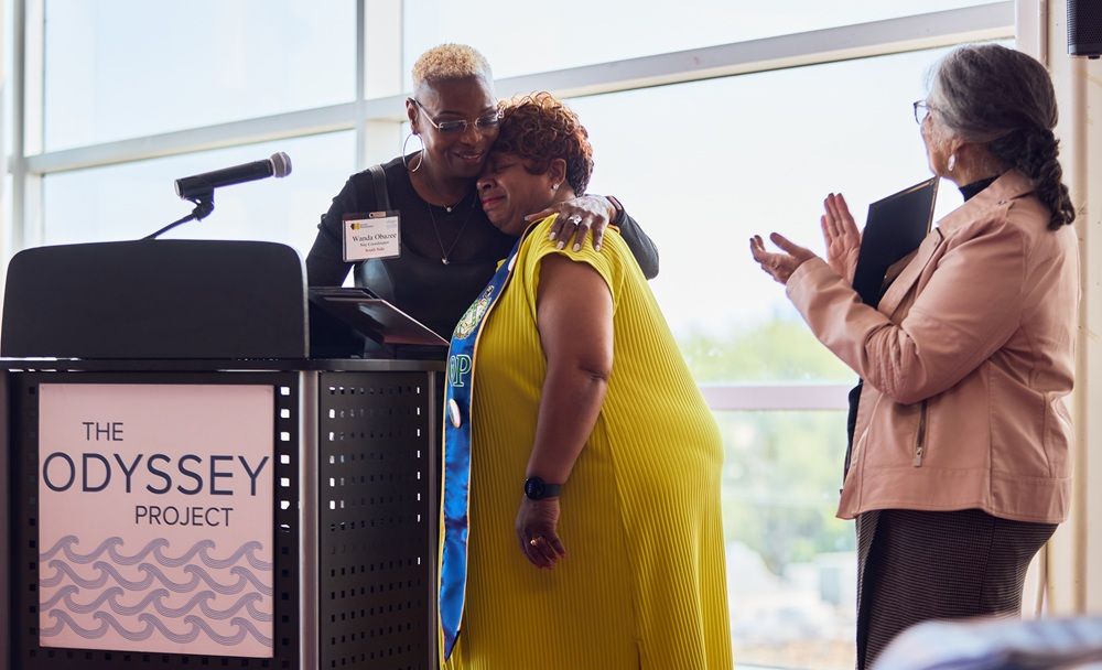 A woman hugs another woman while a third person applauds near a podium with the words "The Odyssey Project"