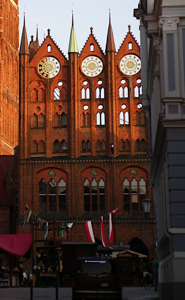 Hochform. Farbe. Im Stil der norddeutschen Backsteingotik errichtetes Rathaus, aus einer Nebenstraße fotografiert. Vor dem Gebäude stehen Verkaufsbuden und hängt eine Girlande mit Fahnen. 