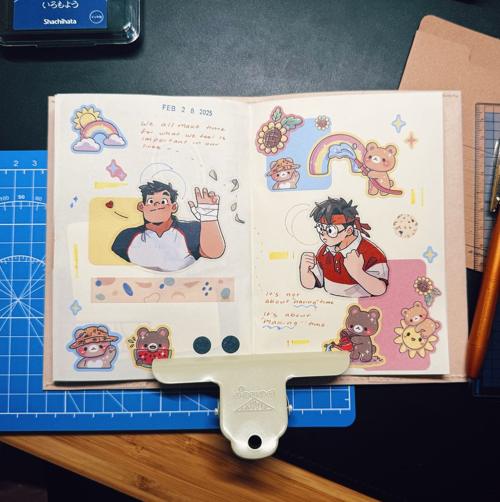 An open journal spread on a desk with a blue cutting mat beneath it. The pages are decorated with colorful stickers, illustrations, and handwritten text. The left page features a drawing of a muscular man in a sports outfit waving, along with bear-themed stickers and a quote in orange ink: “We all make time for what we feel is important in our lives.” The right page showcases a drawing of a boy in glasses and a red headband in a determined pose, surrounded by more bear-themed stickers, rainbows, flowers, and another handwritten quote: “It’s not about having time, it’s about making time.” A large metal clip is holding the pages open. The workspace includes a ruler, an orange pen, and a stamp pad.