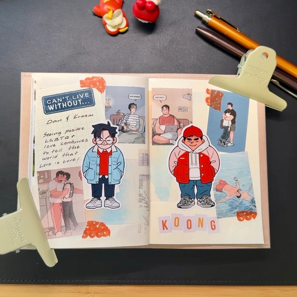 An open journal spread decorated with colorful stickers and cutouts featuring characters Dan and Kaam from a webcomic by the artist Koong. The left page includes a note that reads: “CAN’T LIVE WITHOUT… Dan & Kaam. Seeing positive LGBTQ+ love continues to tell the world that Love is Love!” The page is decorated with orange translucent washi tape dots, and cartoon versions of Dan and Kaam in casual outfits. The right page includes more illustrations of the couple in various moments—cuddling, swimming, and relaxing at home. The word “KOONG” is spelled out in block letters at the bottom. The journal is held open by pale yellow clips, and there are pens and small figurines on the desk above it.