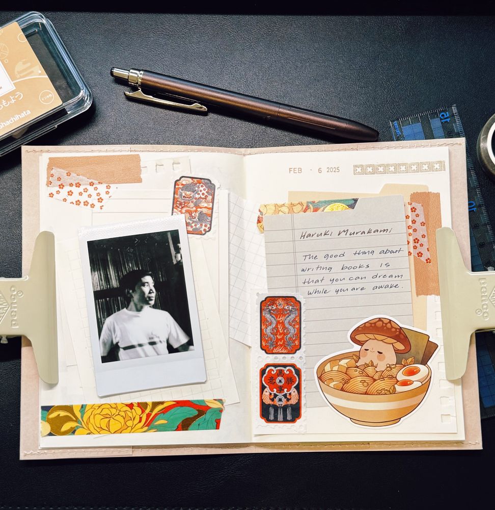 A neatly arranged journal spread features a vintage aesthetic with a mix of decorative elements. The open journal is secured with two metal clips on either side. On the left page, a black-and-white instant photo of Haruki Murakami is attached [image credit Paris Review], surrounded by patterned paper, washi tape, and small decorative stickers. The right page contains a handwritten quote attributed to Murakami: “The good thing about writing books is that you can dream while you are awake.” Below the quote, there is a cute sticker of a bowl of ramen with a smiling mushroom character. The spread incorporates warm colors, intricate patterns, and layered textures. Surrounding the journal are a brown pen, a transparent ink pad container, a ruler, and a black leather desk mat. The date “Feb 6 2025” is stamped at the top of the right page. 