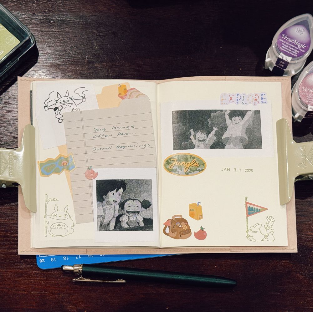 A journal spread with a nostalgic and creative theme featuring images and illustrations from the animated film My Neighbor Totoro. The left page includes a small lined paper with a handwritten quote, “Big things often have… Small beginnings,” along with black-and-white printed images of characters from the movie. Surrounding decorations include washi tape, stickers, and Totoro illustrations. The right page contains additional printed images, stickers with words like “Jungle” and “Explore,” and stamps of rabbits, a flag, and a juice box. The journal is held open with large clips on both sides. A green pen and a green ink pad are placed below the journal, with ink pads visible in the background.