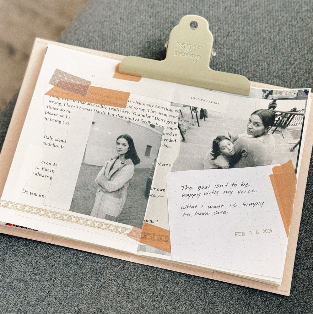 This image features a collage of various paper elements, including book pages, black-and-white photographs, and handwritten notes. The book page has text partially visible, with the name “Jhumpa Lahiri” appearing at the top. One black-and-white photograph depicts Jhumpa Lahiri standing with arms crossed, while another captures Jhumpa Lahiri sitting on stairs with a child, both looking back towards the camera. There are pieces of decorative tape securing the items. A handwritten note on a small, dotted paper reads:

“The goal isn’t to be happy with my voice. What I want is simply to have one.”

The note is dated “FEB 16 2025” and is attached with an orange piece of tape. The clipboard clip is labeled “Penco,” and the entire setup rests on a soft gray fabric surface, creating a cozy and artistic aesthetic.