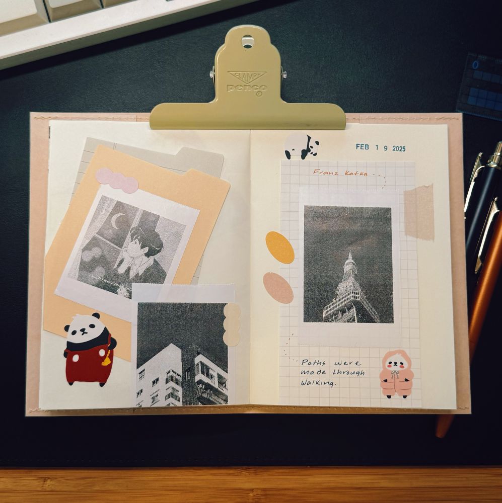 A neatly arranged open journal spread on a black desk, held open with a beige-colored Penco clip. The journal pages contain black-and-white printed images, handwritten text, and decorative stickers. The left page features a grayscale anime-style illustration of a person looking out of a window, a photo of a building, and a small panda sticker wearing red clothes. The right page includes a black-and-white image of Tokyo Tower, handwritten text mentioning “Franz Kafka,” and the quote “Paths were made through walking.” There are also pastel-colored paper cutouts and a small sticker of a hooded animal figure. A mechanical keyboard, pens, and a ruler are visible around the journal.
