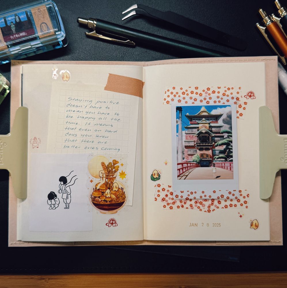 A well-organized journal spread featuring a soft pink notebook held open by two beige clips. The left page contains a handwritten note on grid paper taped to the page with brown washi tape. The note reads: “Staying positive doesn’t have to mean you have to be happy all the time. It means that even on hard days you know that there are better ones coming.” Below the note is a small illustration of two characters from Spirited Away, alongside a colorful sticker depicting a bowl of food with tiny soot sprites. The right page features an instant film photo of the bathhouse from Spirited Away, surrounded by decorative red floral stickers and small character stickers from the film. The date “JAN 28 2025” is stamped at the bottom. Surrounding the notebook are stationery items, including pens, tweezers, and a small plastic container. The scene is lit with warm lighting, creating a cozy and creative workspace ambiance.