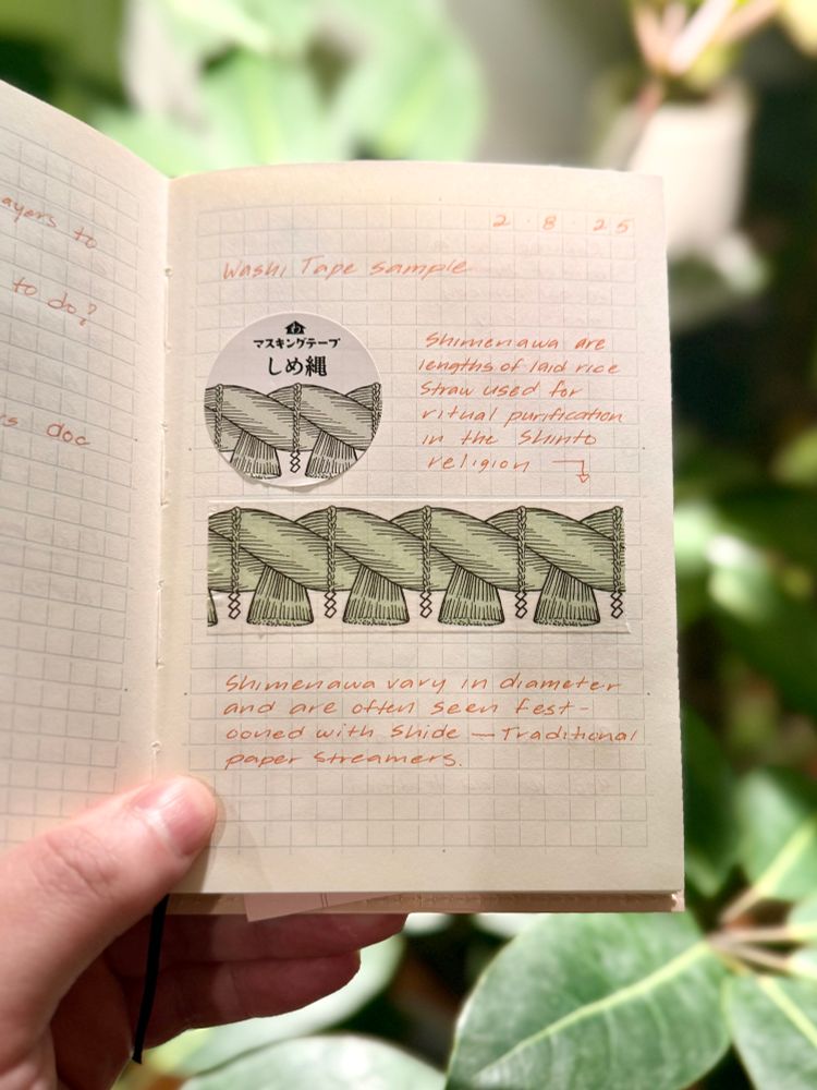 A person holds an open notebook with a grid-patterned page, featuring a washi tape sample with a “shimenawa” (a traditional Japanese rope used in Shinto rituals) design. The tape is adhered to the page, along with a circular label containing Japanese text. There are handwritten notes in red ink explaining that “shimenawa are lengths of laid rice straw used for ritual purification in the Shinto religion” and that they “vary in diameter and are often seen festooned with shide—traditional paper streamers.” The background consists of blurred green leaves, indicating an outdoor or natural setting.