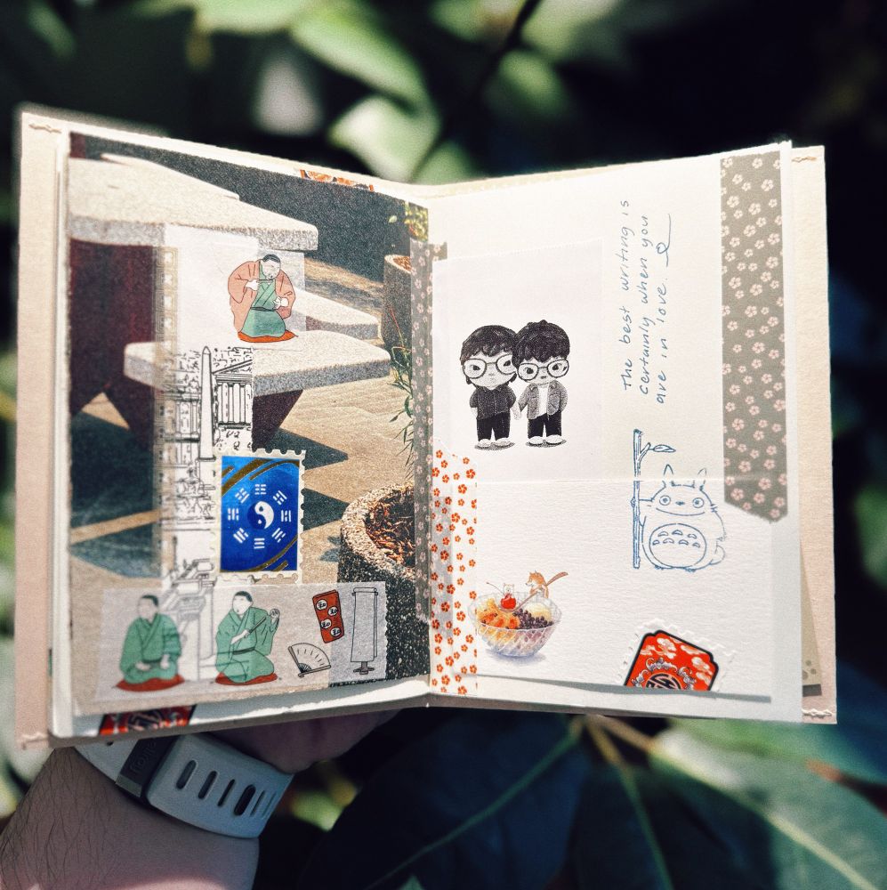  A person holds an open scrapbook outdoors with green foliage in the background. The left page contains a collage featuring a photograph of concrete stairs with a small illustration of a person sitting on them, along with various stickers and cutouts, including Japanese postage stamps, traditional Japanese figures in green robes. The right page has a hand-drawn illustration of two cartoonish people wearing glasses, a blue ink drawing of Totoro holding a bamboo stick, a small sticker of a dessert bowl with a tiny Shiba, and a handwritten note reading, “The best writing is certainly when you are in love.” The pages are decorated with patterned washi tape and have a textured, handmade feel.