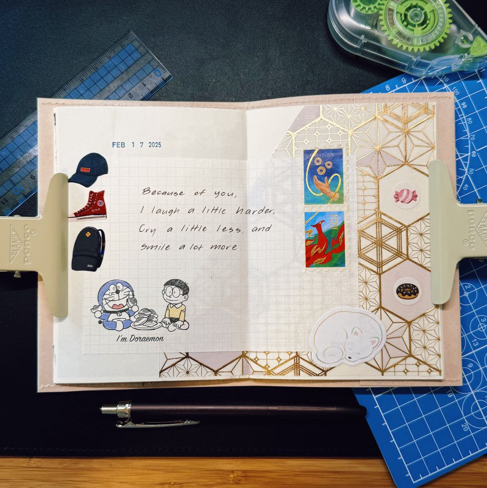 A creative journal spread open on a desk, held down with two beige clips. The left page contains a handwritten quote: “Because of you, I laugh a little harder, cry a little less, and smile a lot more.” It is decorated with small stickers, including a blue cap, a red sneaker, a blue backpack, and a “I’m Doraemon” illustration. The right page has gold geometric designs, small hexagonal stickers with illustrations, and colorful abstract artwork. Surrounding the journal are crafting tools, including a ruler, a mechanical pen, and a gear-pattern tape cutter. The workspace is neatly arranged on a black surface with a blue cutting mat partially visible.