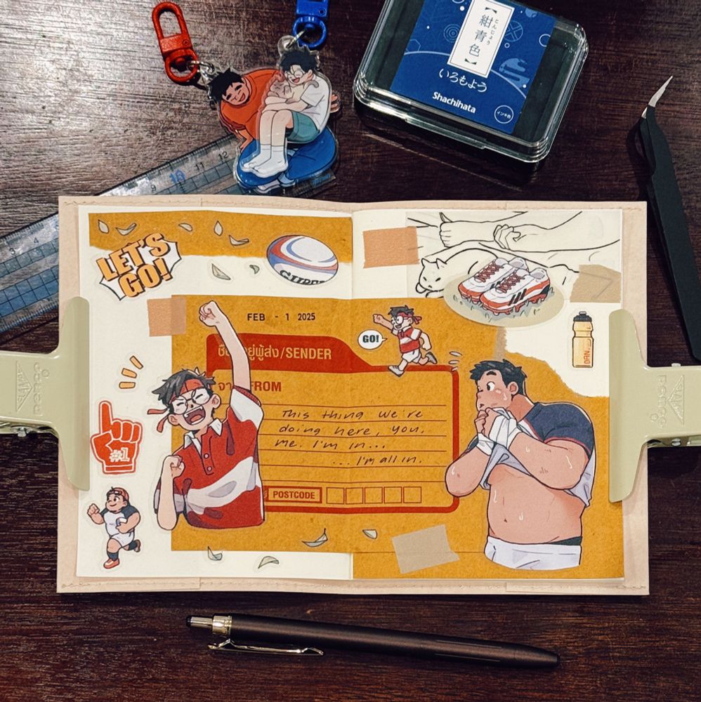 A well-organized journal spread on a wooden desk features a rugby-themed collage with various stickers, drawings, and handwritten notes. The spread has a warm orange and beige color scheme with illustrations of animated characters in rugby attire, a rugby ball, and motivational phrases like ‘LET’S GO!’ taped onto the page. A notable handwritten note reads: ‘This thing we’re doing here, you, me. I’m in… I’m all in.’ Surrounding the journal are stationery items, including a black pen, a ruler, a craft knife, and a blue ink pad labeled ‘Shachihata.’ Two acrylic keychains with anime-style characters are clipped to a zipper pull. The journal is held open with beige paper clamps, and small decorative elements like washi tape and cut-out illustrations enhance the composition.