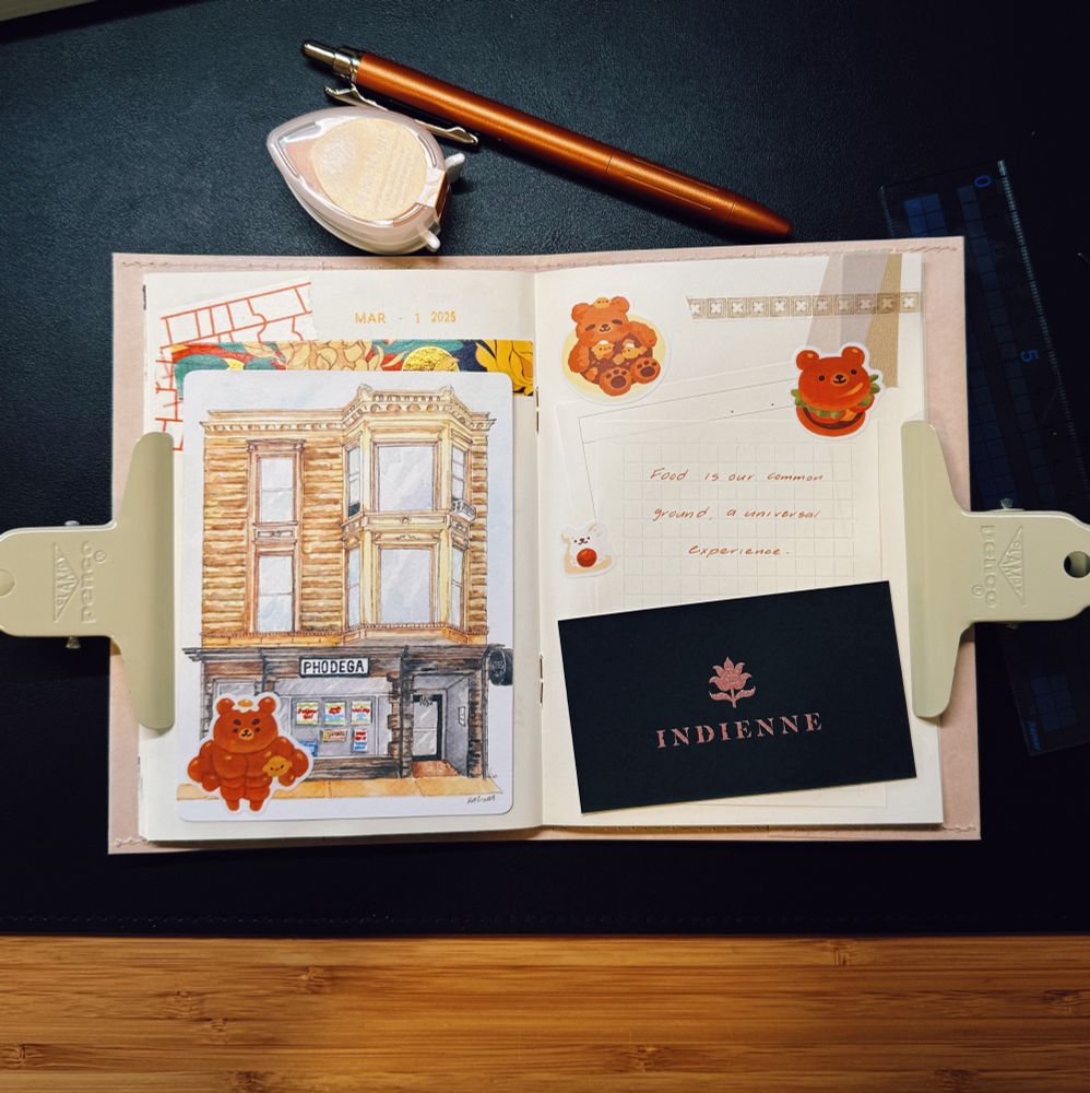 The image shows an open journal spread with various decorative elements and stationery on a black surface. The left page features a watercolor or ink illustration of a two-story brick building with a sign reading “PHODEGA,” accompanied by bear stickers. The right page contains handwritten text in orange ink that reads, “Food is our common ground, a universal experience,” along with grid paper, bear-themed stickers, and a black business card with the word “INDIENNE” and a pink leaf design. The journal is held open with two beige clips. Surrounding the journal are an orange pen, a VersaMagic ink pad, and a ruler, all placed neatly on the desk. The scene has a warm and artistic feel, showcasing a mix of journaling, sketching, and creative organization.