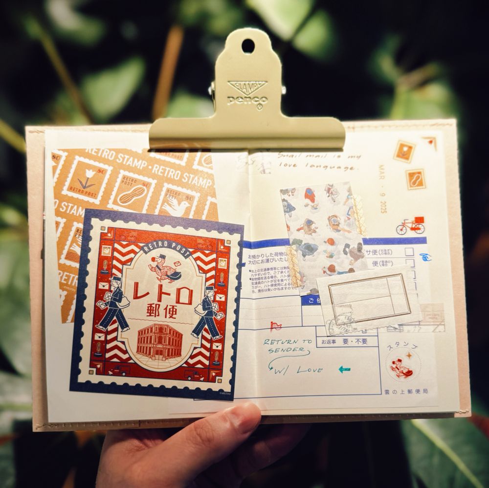A person is holding an open journal or scrapbook with a metal clip labeled “Penco.” The pages contain a collage of vintage-inspired postage-themed designs, stamps, stickers, and handwritten notes. The left page features a large, retro-style postage stamp with Japanese text and an illustration of a bird carrying a letter. The right page includes layered elements such as airmail-inspired stickers, a small piece of decorative washi tape with illustrations of people, and a handwritten message that reads, “Snail mail is my love language.” Another handwritten note in blue ink says, “RETURN TO SENDER w/ Love.” The background consists of blurred green foliage, creating a warm and nostalgic atmosphere.