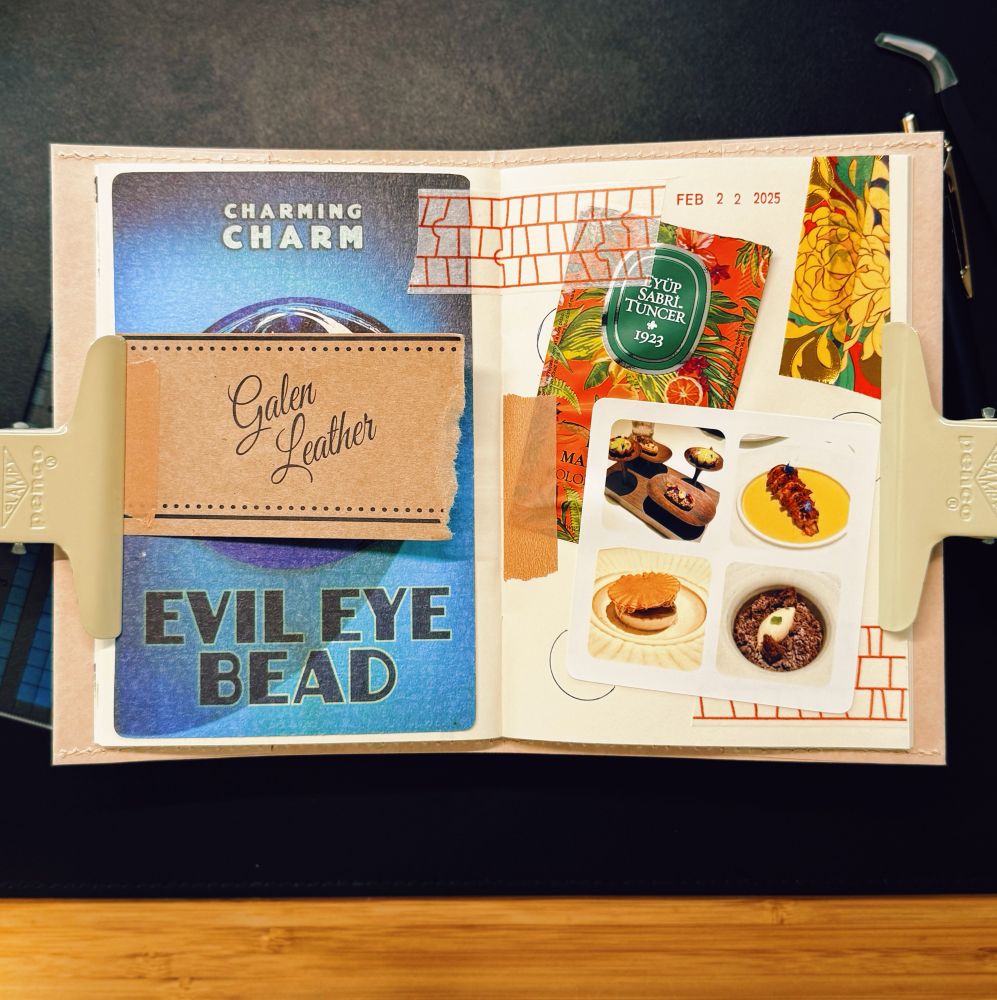 The image shows an open journal spread with a collage-style layout, held in place by two beige clips on either side. The left page features a blue card with the words “EVIL EYE BEAD” and “CHARMING CHARM,” along with a brown tag labeled “Galen Leather,” attached with washi tape. The right page contains a mix of decorative elements, including a vintage-style floral print, a green sticker labeled “Eyüp Sabri Tuncer 1923,” and a small collage of food photographs showing various plated dishes. Hand-stamped elements such as a red brick pattern and a date stamp reading “FEB 22 2025” are also present. The background is a dark surface, possibly a desk, with a pen partially visible at the top right. The journal appears to be a travel or memory-keeping notebook with a mix of textures and printed materials.