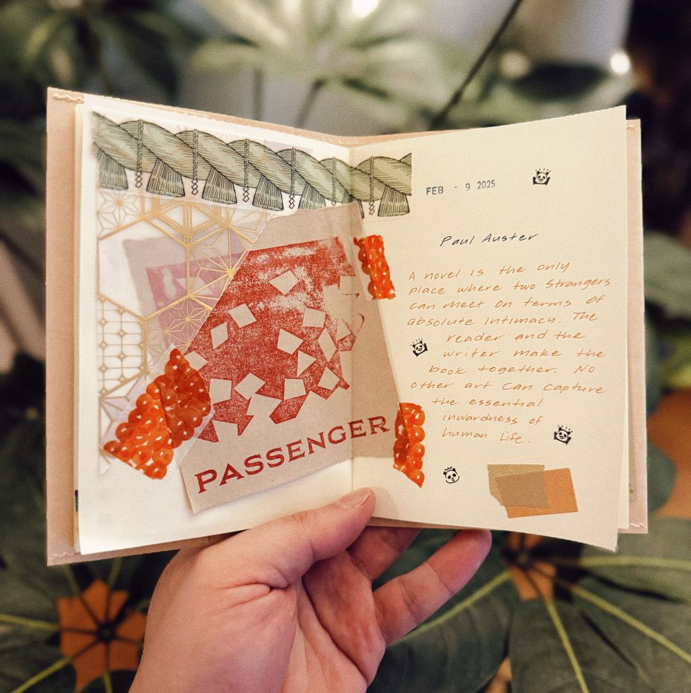 A person holds an open handmade journal with a mixed-media collage spread. The left page features layered decorative elements, including geometric patterns, textured paper, and a stamped red print with the word “PASSENGER.” A strip of washi tape depicting salmon roe is placed on the page, adding a textured visual effect. The right page contains a typed date, “FEB · 9 2025,” and a handwritten quote by Paul Auster in orange ink:
“A novel is the only place where two strangers can meet on terms of absolute intimacy. The reader and the writer make the book together. No other art can capture the essential inwardness of human life.”
Small illustrated stamps, washi tape, and color swatches add to the artistic composition. The background features a large green plant with broad leaves.