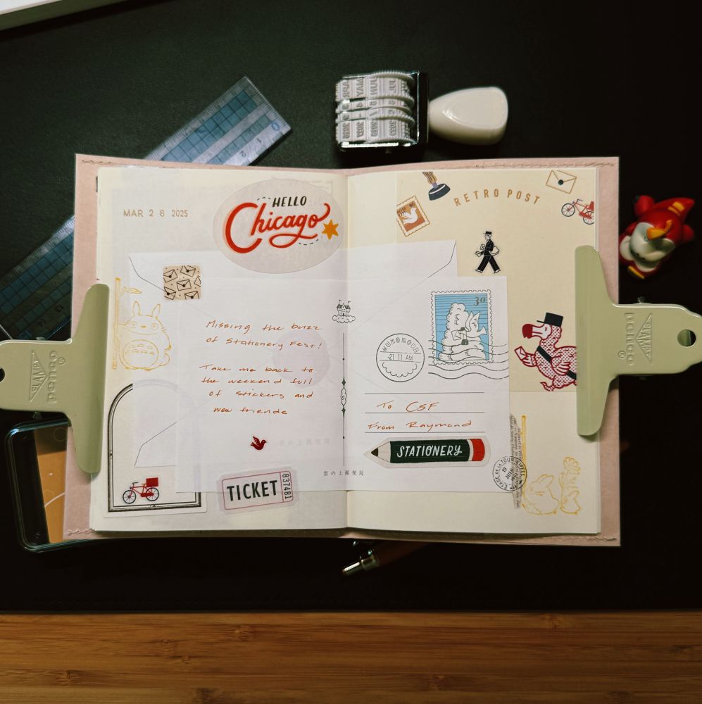 An open journal decorated with colorful stationery-themed stickers and stamps, featuring a large “Hello Chicago” sticker dated March 28, 2025. A handwritten note reads, “Missing the buzz of stationery fest! Take me back to the weekend full of stickers and new friends.” The pages are clipped open with beige metal clips. Surrounding the journal are items including a date stamp, eraser, ruler, and a small red toy figure.