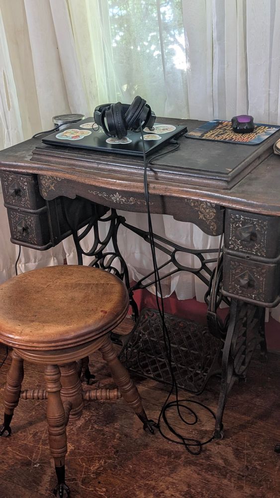Treadle sewing machine with the top closed. In front of it is a piano stool. On top of it is a closed laptop computer,  headphones, and mouse 