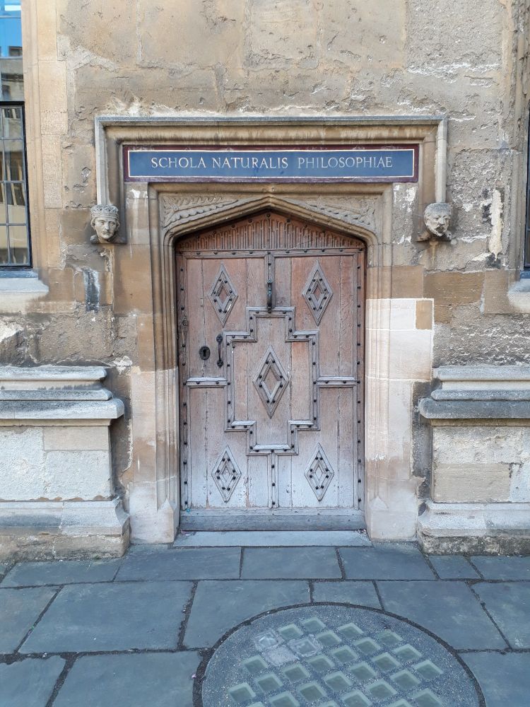An ancient wooden door in an old stone wall, with a blue sign above, lettered in gold "Schola Naturalis Philosophiae"