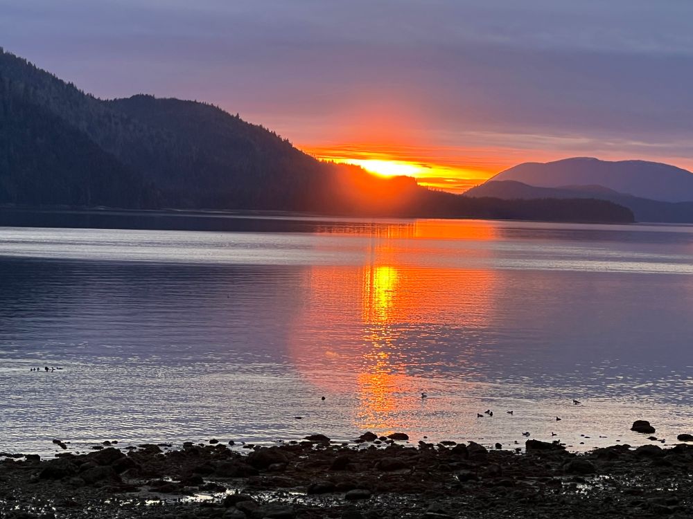 Bright Orange sunset behind island and calm ocean and rocky southeast Alaskan beach. 