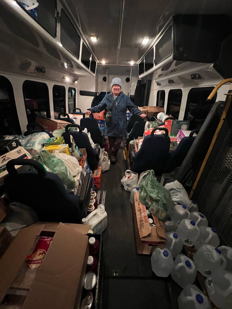 View of inside the bus. Food goods piled on seats and floor. Person is standing in the middle aisle. 