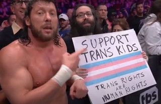AEW pro wrestler Kenny Omega pointing to a sign, held by a fan in the audience of a show, that says “ support trans kids trans rights are human rights “
